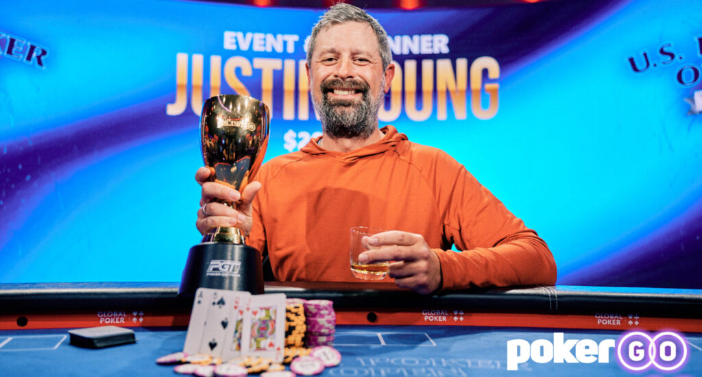 Man with gray beard in an orange shirt holds a gold trophy at a poker tournament, with chips and a drink on the table in front of a blue backdrop showing a prize banner.